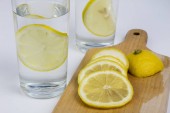 Sklenici studené vody s citronem na bílé kuchyňském stole. Osvěžující nápoj v teplém počasí. Světlé pozadí.