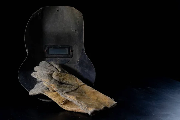 An old mask a protective suit for a welder. Occupational health and safety clothing for production workers on a workshop table. Black background.