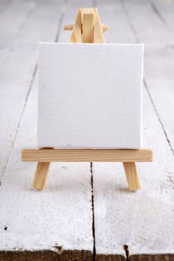Tahta bir masanın üzerinde beyaz, boş bir bez bulunan küçük bir sehpa. Minyatür boya aksesuarları. Hafif arkaplan.
