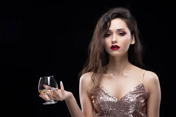Beautiful glamorous brunette girl wearing a sparkling dress sensually holds in her hand a glass of whiskey with ice. Shimmering makeup, red lips. Isolated on black. Copy space. Commercial design