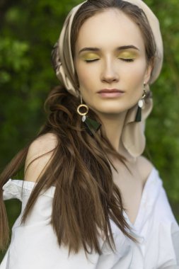 Trendy bir beyaz gömlek, bej pantolon ve bir pareo saçlarını üzerinde giyen güzel şık genç kız açık havada yeşil yaprakları bir arka plan karşı teşkil etmektedir. Ticari ve reklam tasarımı. Kopya alanı