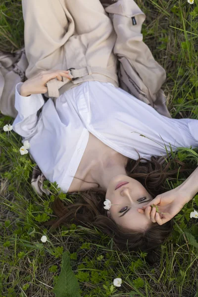 Güzel şık genç kız trendy bir beyaz gömlek, bej jimnastik pantolonu ve bir pareo kır çiçekleri arasında yeşil çimenlerin üzerinde rahatlatıcı yatıyor. Ticari ve reklam tasarımı. Kopya alanı.