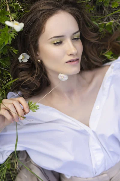 Güzel şık genç kız trendy bir beyaz gömlek, bej jimnastik pantolonu ve bir pareo kır çiçekleri arasında yeşil çimenlerin üzerinde rahatlatıcı yatıyor. Ticari ve reklam tasarımı. Kopya alanı.