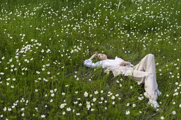 Güzel şık genç kız trendy bir beyaz gömlek, bej jimnastik pantolonu ve bir pareo kır çiçekleri arasında yeşil çimenlerin üzerinde rahatlatıcı yatıyor. Ticari ve reklam tasarımı. Kopya alanı.