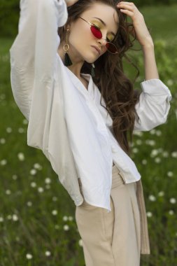 Trendy bir beyaz gömlek, bej jimnastik pantolonu ve kırmızı güneş gözlüğü giyen güzel şık genç kız açık havada beyaz kır çiçekleri tam çayır teşkil etmektedir. Ticari ve reklam tasarımı. Kopya alanı
