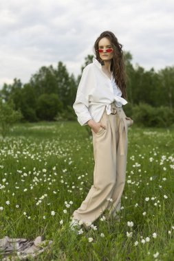 Trendy bir beyaz gömlek, bej jimnastik pantolonu ve kırmızı güneş gözlüğü giyen güzel şık genç kız açık havada beyaz kır çiçekleri tam çayır teşkil etmektedir. Ticari ve reklam tasarımı. Kopya alanı