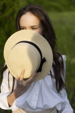 Trendy bir beyaz gömlek, bej pantolon ve açık havada yeşil yaprakları bir çerçevede pozlar gülümseyen hasır şapka şapka giyen güzel şık genç kız. Reklam ve moda tasarımı. Kopya alanı.