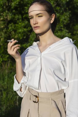 Trendy giyen güzel şık yasaktır kız gömlek, bej pantolon beyaz ve açık havada güneş ışığı tutar içinde peruk altında yeşil çayır onun elinde bir sigara kafes. Reklam tasarımı. Kopya alanı.