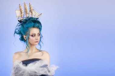 Sensual young girl with naked shoulders and painted blue hair stowed in an artistic hairdo with a toy sailboat in her hair, covering her chest with blue and white veils. Blue background. Copy space clipart
