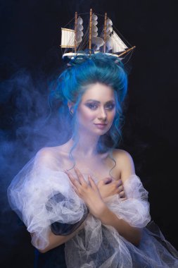 Sensual young girl with naked shoulders and painted blue hair stowed in an artistic hairdo with a toy sailboat in her hair, covering her chest with blue and white veils in a scenic smoke. Copy space clipart