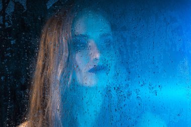 Sanat stil portre bir pencere cam arkasında bir yalnız güzel üzgün model kız mavi tonları, yağmur bitti düşer. Sanatsal bulanıklık modeli yüzünde. Kavramsal, ticari ve şık. Kopya alanı