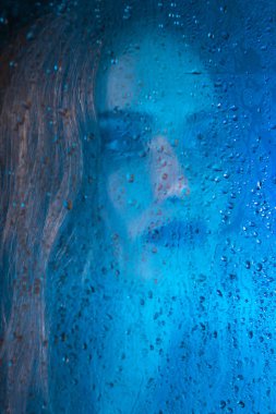 Sanat stil portre bir pencere cam arkasında bir yalnız güzel üzgün model kız mavi tonları, yağmur bitti düşer. Kavramsal, ticari ve moda tasarımı. Kopya alanı.