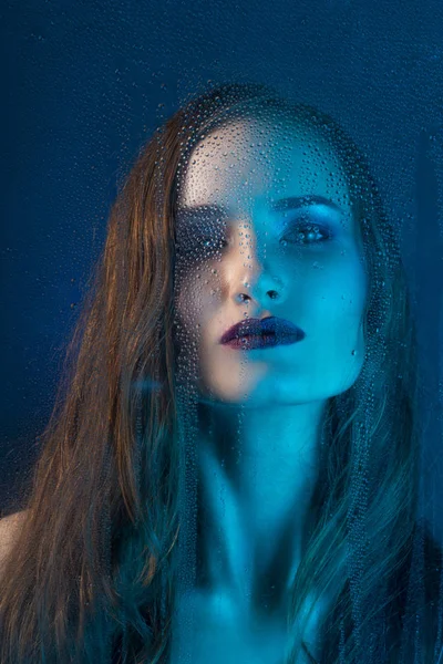 Art style portrait in blue tones of a lonely beautiful sad model girl behind a window glass, over which rain drops down. Conceptual, commercial and fashionable design. Copy space.