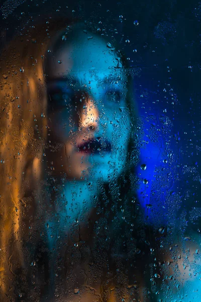 Art style portrait in blue tones of a lonely beautiful sad model girl behind a window glass, over which rain drops down. Artistic blur on model face. Conceptual, commercial and fashionable. Copy space
