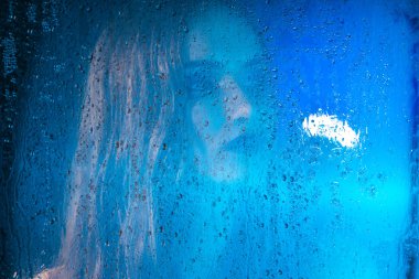 Sanat stil portre bir pencere cam arkasında bir yalnız güzel üzgün model kız mavi tonları, yağmur bitti düşer. Sanatsal bulanıklık modeli yüzünde. Kavramsal, ticari ve şık. Kopya alanı