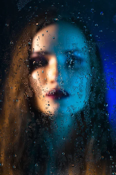 Art style portrait in blue tones of a lonely beautiful sad model girl behind a window glass, over which rain drops down. Artistic blur on model face. Conceptual, commercial and fashionable. Copy space