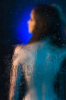 Camın arkasında duran güzel bir ince çıplak kız bulanık siluet su damlaları ile kaplı. Sanatsal ve kavramsal fotoğraf mavi tonlarında. Kopya alanı.