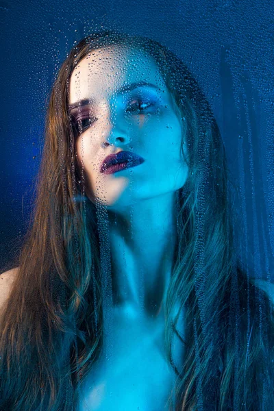 Art style portrait in blue tones of a lonely beautiful sad model girl behind a window glass, over which rain drops down. Conceptual, commercial and fashionable design. Copy space.