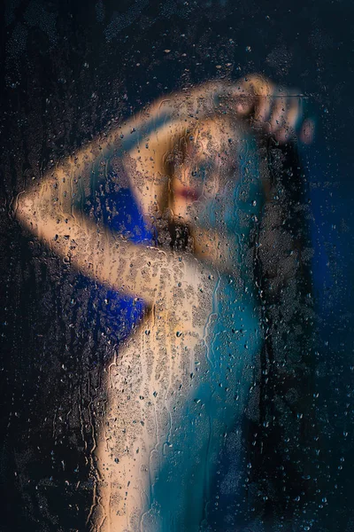 ガラスの後ろに立って美しい細い裸の女の子のぼやけたシルエットは水滴で覆われています ブルーの色調で芸術的 概念的な写真 コピー スペース — ストック写真
