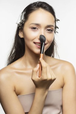 Güzel genç esmer kız modeli ile çıplak omuz ve saç kulpları, Islak saçla beyaz arka plan üzerinde yüzünde makyaj fırçası ile koyar. Sağlıklı temiz cilt. Reklam ve ticari tasarım