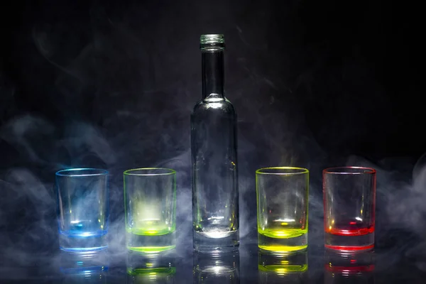 Five multicolored empty shot glasses and small bottle placed symmetrically on a black background in a theatrical smoke. Conceptual, commercial and advertising photo. Copy space.