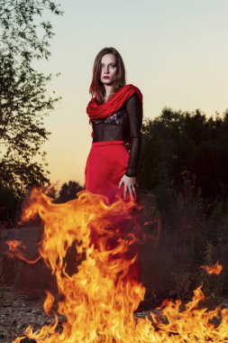 Kırmızı uzun etek ve kırmızı şal giyen güzel moda modeli kız'nde günbatımı büyük ateşin yanında duruyor. Kavramsal, şık, modern ve reklam tasarım.
