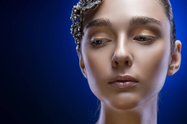 Güzel genç esmer model kızla çıplak omuz takı diadem mavi-siyah bir degrade arka plan üzerinde giyiyor. Altın makyaj. Sağlıklı temiz cilt. Uzay kopyalayın. Reklam ve konsept hazırlama.