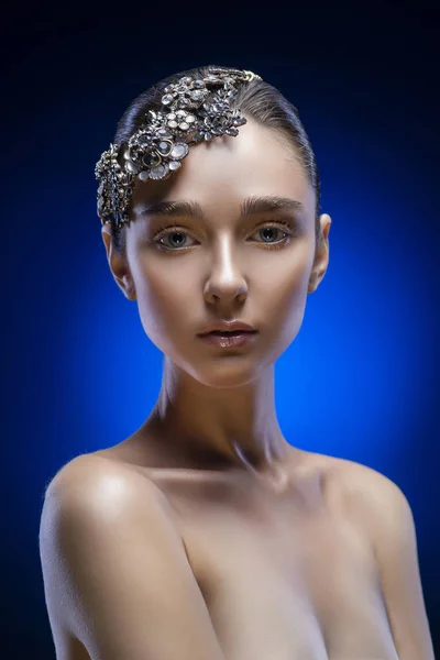 Beautiful young brunette model girl with naked shoulders wearing jewelry diadem on a blue-black gradient background. Golden make-up. Healthy clean skin. Copy space. Advertising and conceptual design.