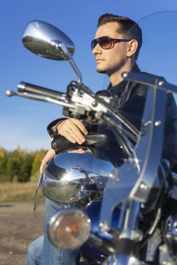 Bir siyah deri ceket, güneş gözlüğü ve kot giyen genç adam açık mavi gökyüzü arka plan üzerinde bir dağda bir motosiklet oturur. Yaşam biçimi, seyahat. Uzay kopyalayın. Reklam ve ticari tasarım.