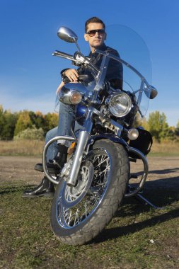 Bir siyah deri ceket, güneş gözlüğü ve kot giyen genç adam açık mavi gökyüzü arka plan üzerinde bir dağda bir motosiklet oturur. Yaşam biçimi, seyahat. Uzay kopyalayın. Reklam ve ticari tasarım.