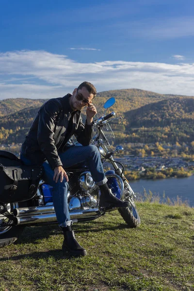 Young man wearing a black leather jacket, sunglasses and jeans sits outdoor on a motorcycle, resting on a mountain above the river. Lifestyle, travel. Copy space. Advertising and commercial design.