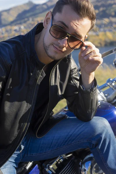 Young man wearing a black leather jacket, sunglasses and jeans sits outdoor on a motorcycle, resting on a mountain above the river. Lifestyle, travel. Copy space. Advertising and commercial design.