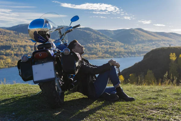 Young man wearing a black leather jacket and jeans sits outdoor on a ground at a motorcycle, resting on a mountain above the river. Lifestyle, travel. Copy space. Advertising design.