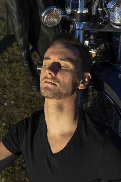 Young man wearing a black leather jacket and jeans sits outdoor on a ground at a motorcycle, resting on a mountain above the river. Lifestyle, travel. Copy space. Advertising design.