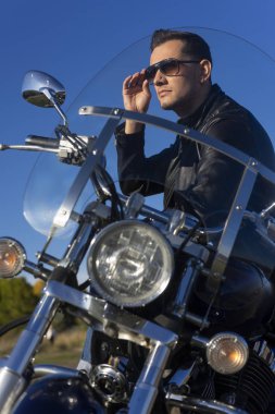 Bir siyah deri ceket, güneş gözlüğü ve kot giyen genç adam açık mavi gökyüzü arka plan üzerinde bir dağda bir motosiklet oturur. Yaşam biçimi, seyahat. Uzay kopyalayın. Reklam ve ticari tasarım.