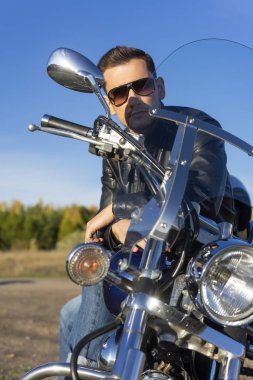 Bir siyah deri ceket, güneş gözlüğü ve kot giyen genç adam açık mavi gökyüzü arka plan üzerinde bir dağda bir motosiklet oturur. Yaşam biçimi, seyahat. Uzay kopyalayın. Reklam ve ticari tasarım.