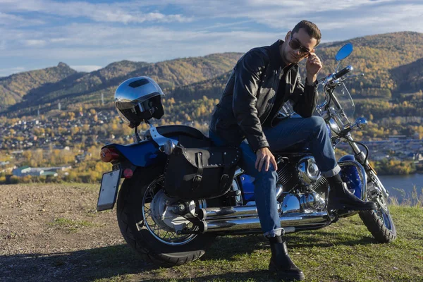 Young man wearing a black leather jacket, sunglasses and jeans sits outdoor on a motorcycle, resting on a mountain above the river. Lifestyle, travel. Copy space. Advertising and commercial design.