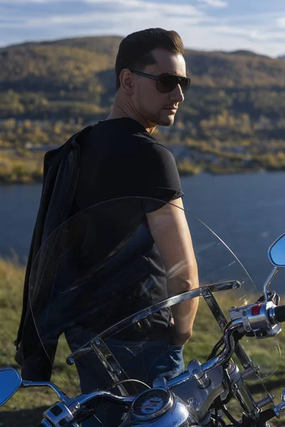 Young man wearing a black leather jacket, sunglasses and jeans stays outdoor at a motorcycle, resting on a mountain above the river. Lifestyle, travel. Copy space. Advertising and commercial design
