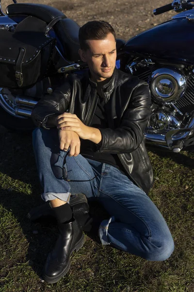 Young man wearing a black leather jacket and jeans sits outdoor on a ground at a motorcycle, resting on a mountain above the river. Lifestyle, travel. Copy space. Advertising design.