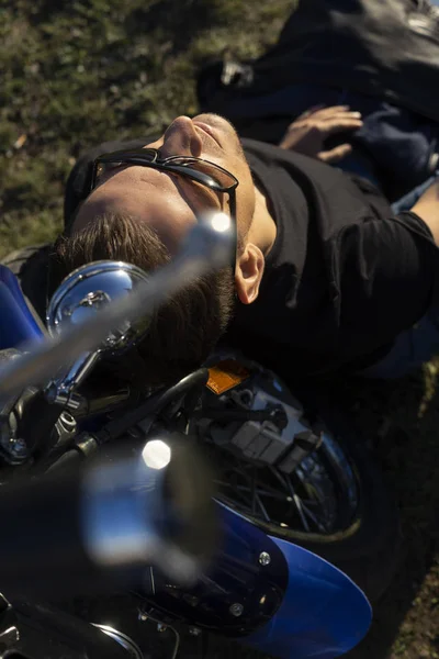 Young man wearing a black leather jacket, sunglasses and jeans sits outdoor on a ground at a motorcycle, resting on a mountain above the river. Lifestyle, travel. Copy space. Advertising design.