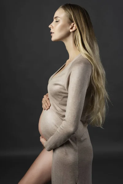 Güzel hamile genç kadın, bir iç çamaşırı ve örme elbise, geri alınan ve çıplak karnı Open'da usulca giymiş karnı elleri dokunur. Gri arka plan üzerinde profil fotoğrafı. Kopya alanı.