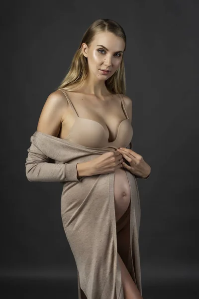 Beautiful pregnant young woman, wearing a lingerie and knitted beige dress, bares her shoulders and holds the dress on her naked belly. Gray background. Fashion, pregnancy, lifestyle. Copy space.