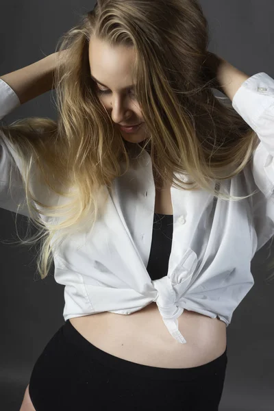 A beautiful smiling pregnant young blonde woman wearing a black maternity underwear and a white unbuttoned shirt, tied on her stomach, touches her hair with her hand. Health, fashion. Copy space.