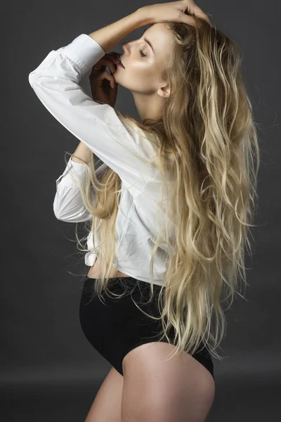 A beautiful smiling pregnant young blonde woman wearing a black maternity underwear and a white unbuttoned shirt, tied on her stomach, touches her hair with her hand. Health, fashion. Copy space.