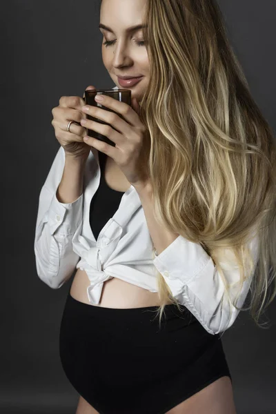 A beautiful smiling pregnant young female blonde wearing a black maternity underwear and a white unbuttoned shirt, tied on her stomach, holds a cup of hot drink in her hands. Copy space.