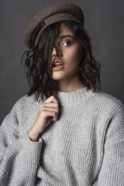 Bir rahat tarzı örgü kazak ve kap giyen güzel korkmuş esmer kız saçları gri arka plan üzerinde eliyle dokunur. Doğal çıplak makyaj. Moda, reklam tasarımı. Kopya alanı