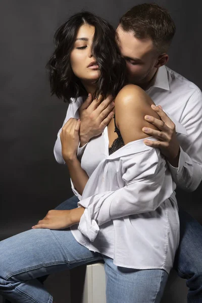 Beautiful couple wearing white shirts and jeans are sitting on a white cube. Man passionately hugs a girl whose shoulder is bare. Casual style. Lifestyle, fashion, commercial design. Gray background.