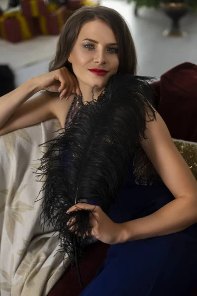 A beautiful slim smiling girl dressed in a long evening dress sits on the couch and holds in her hand a big feather in a festive Christmas interior. New year, lifestyle, fashion design. Copy space.