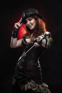 Derin bir yaka bir Victorian tarzı steampunk kostüm ve büyük meme ile şapka giyen güzel gülümseyen Kızıl saçlı cosplayer kız kamışı uzanmış elinde tutar. Kırmızı ve siyah arka plan. Kopya alanı.