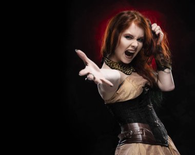 Güzel kızıl saçlı cosplayer kız saçını eliyle Victorian tarzı steampunk elbise çığlıklar, shooks giyen ve elini outstretching. Kırmızı ve siyah arka plan. Kopya alanı.